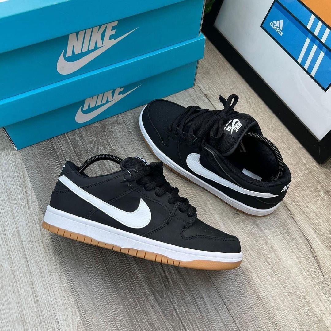 Black Nike Dunk Low sneakers with white swoosh and gum sole next to Nike boxes and Adidas frame.