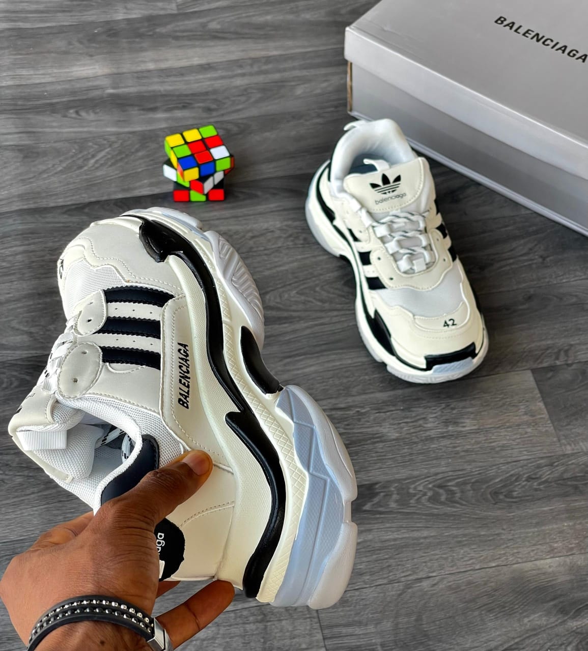 Hand holding white Balenciaga x Adidas Triple S chunky sneaker with second pair and box visible.