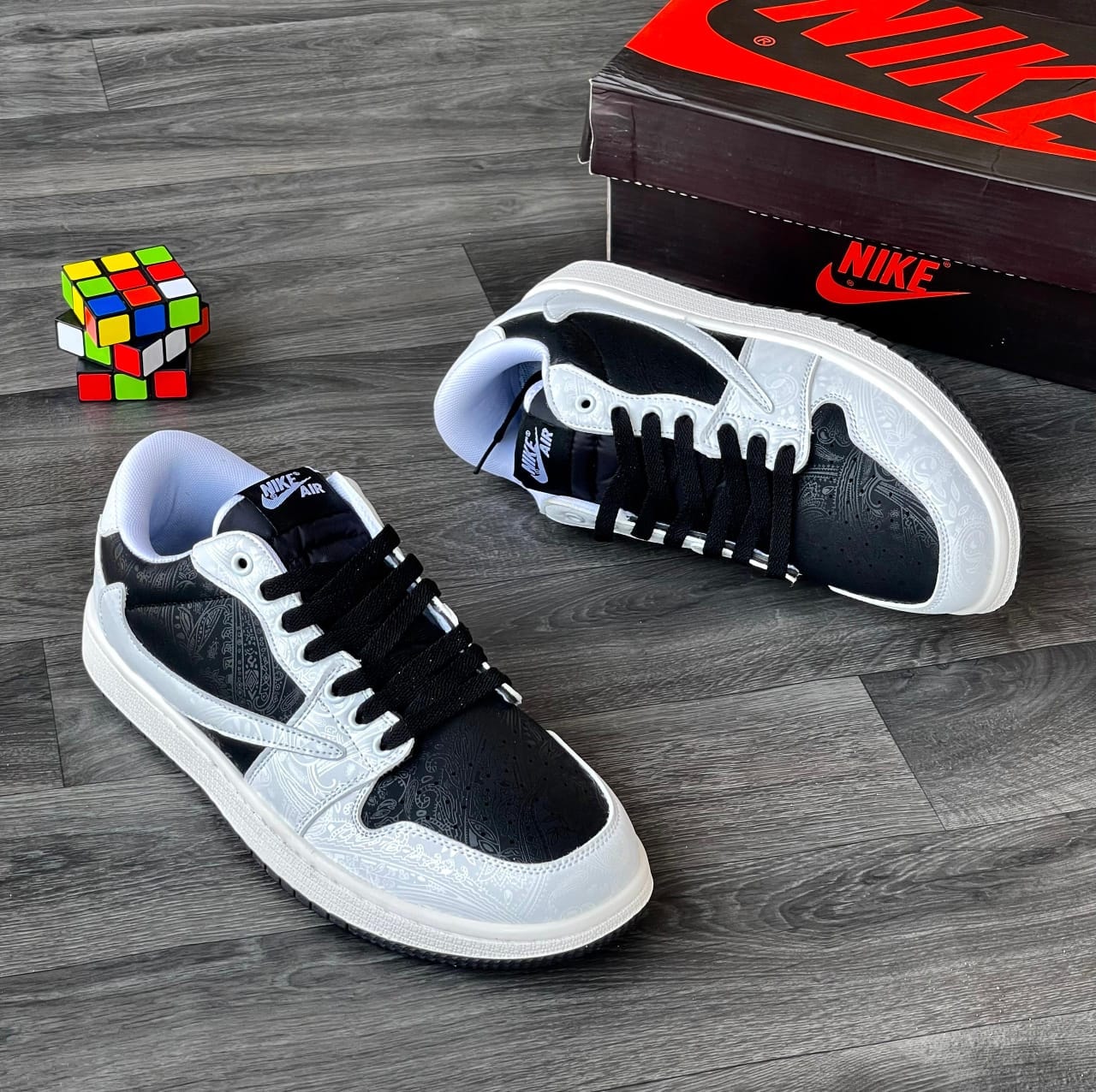 Black and white paisley Nike Air Jordan 1 Low sneakers on wooden floor with Nike box and Rubik's cube.