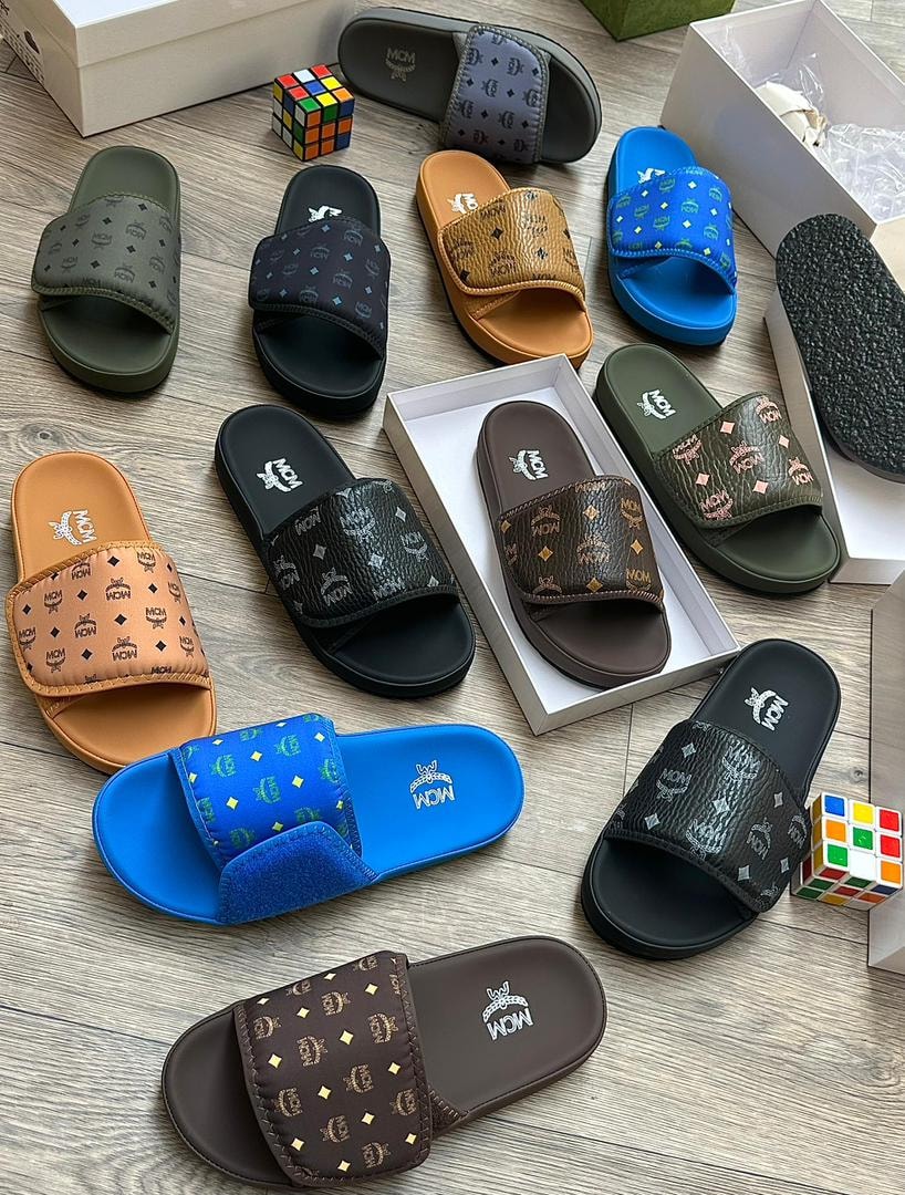 Flat lay showing multiple MCM monogram slides in various colors on wooden surface.