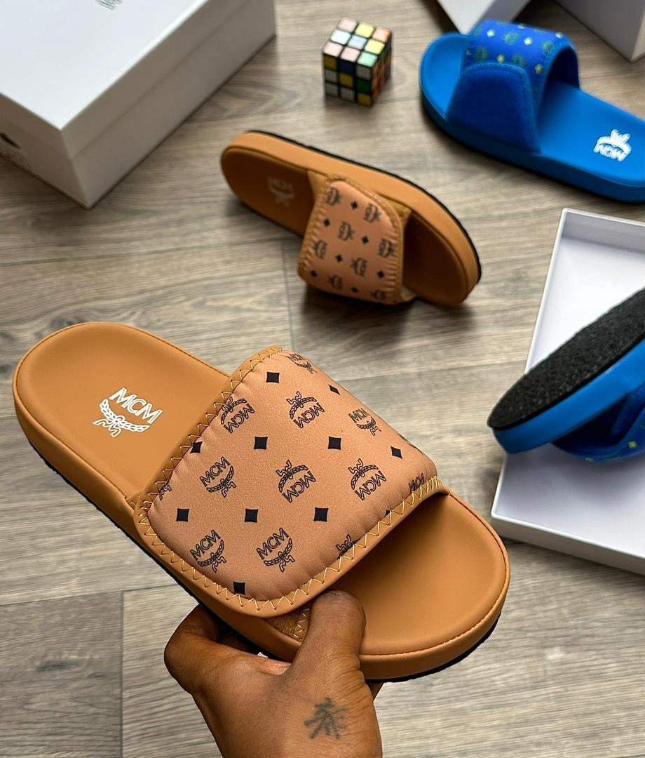 Tan MCM monogram slide with black logos held in hand with background items.