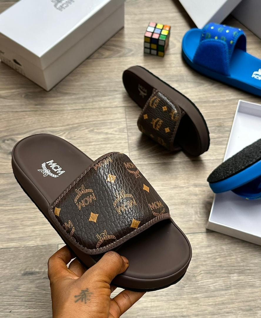 Brown MCM monogram slide with gold logos held in hand on wooden floor.