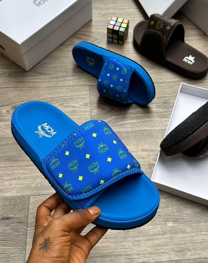 Blue MCM monogram slide with lime green logos held in hand, other slides visible.
