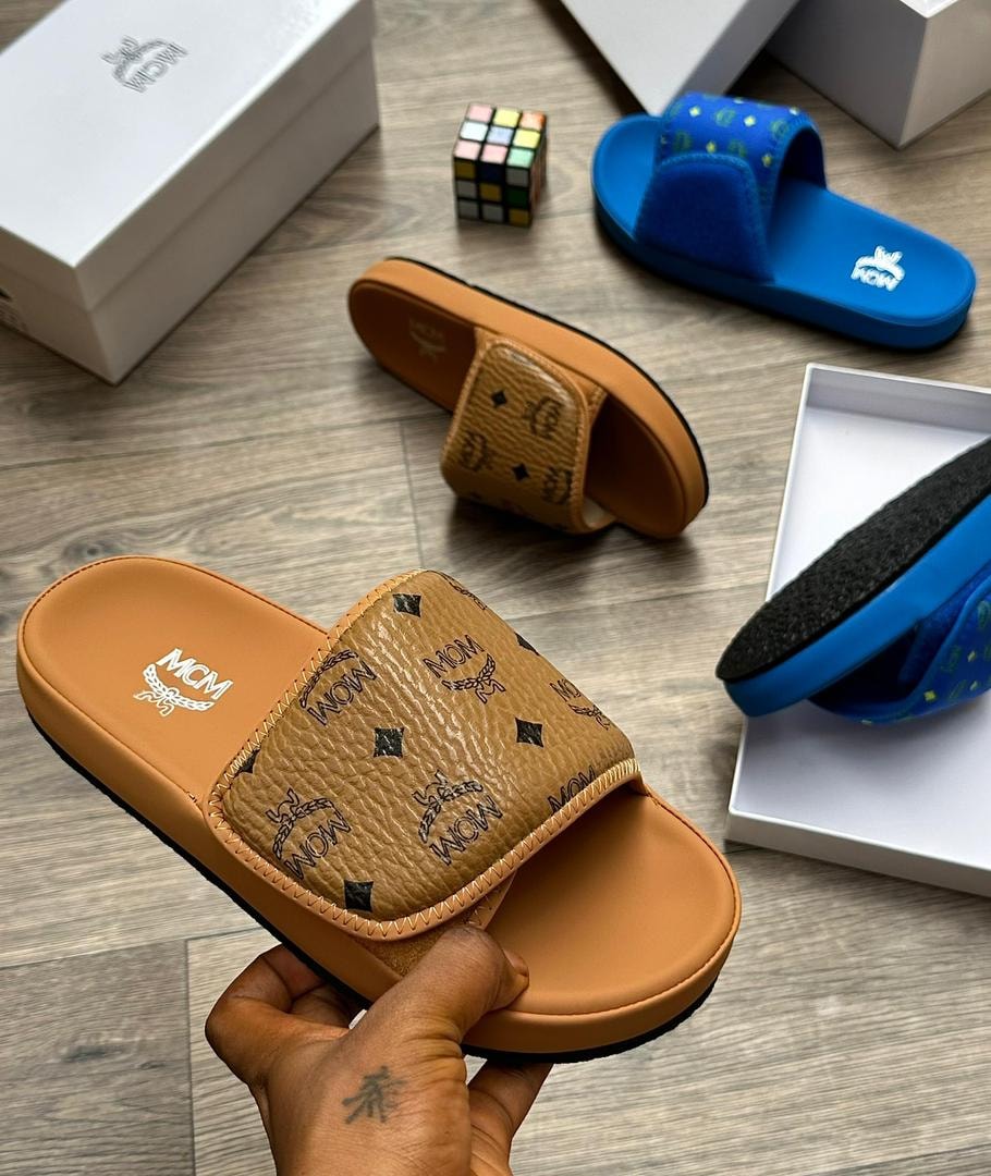 Tan MCM monogram slide with black logos held in hand, blue and other slides in background.
