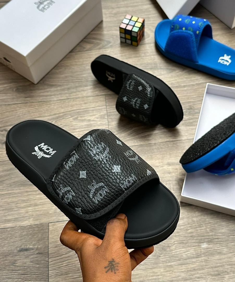 Black MCM monogram slide with white logos held in hand with other colorways visible.