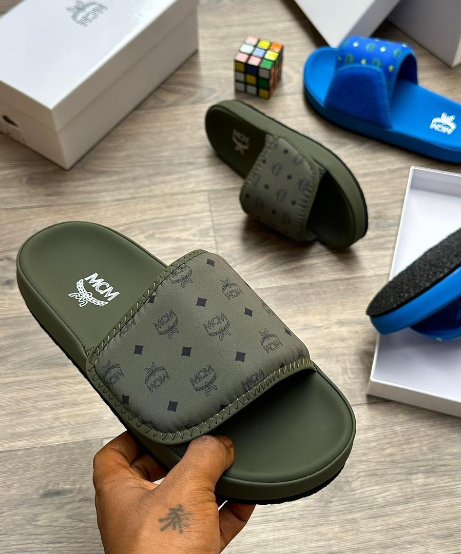 Olive MCM monogram slide with dark logos held in hand on wooden surface with packaging.