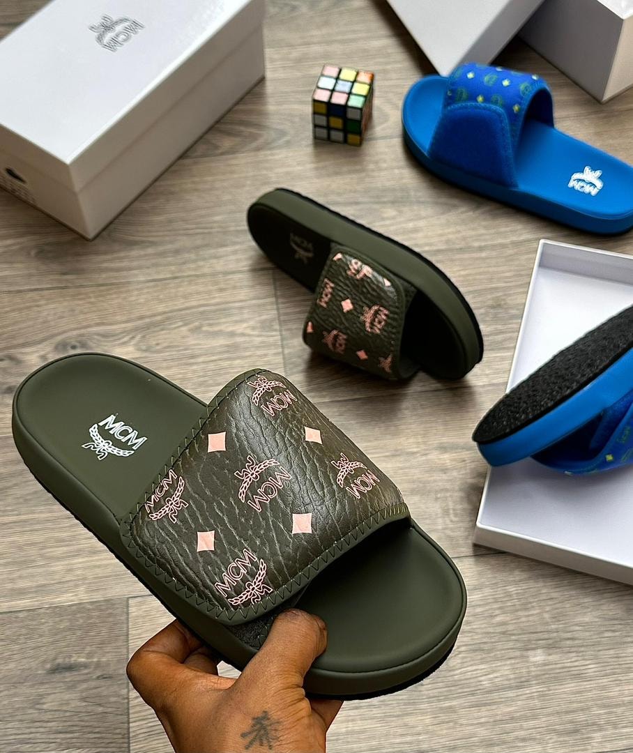 Olive green MCM monogram slide with pink logos held in hand, other slides visible in background.