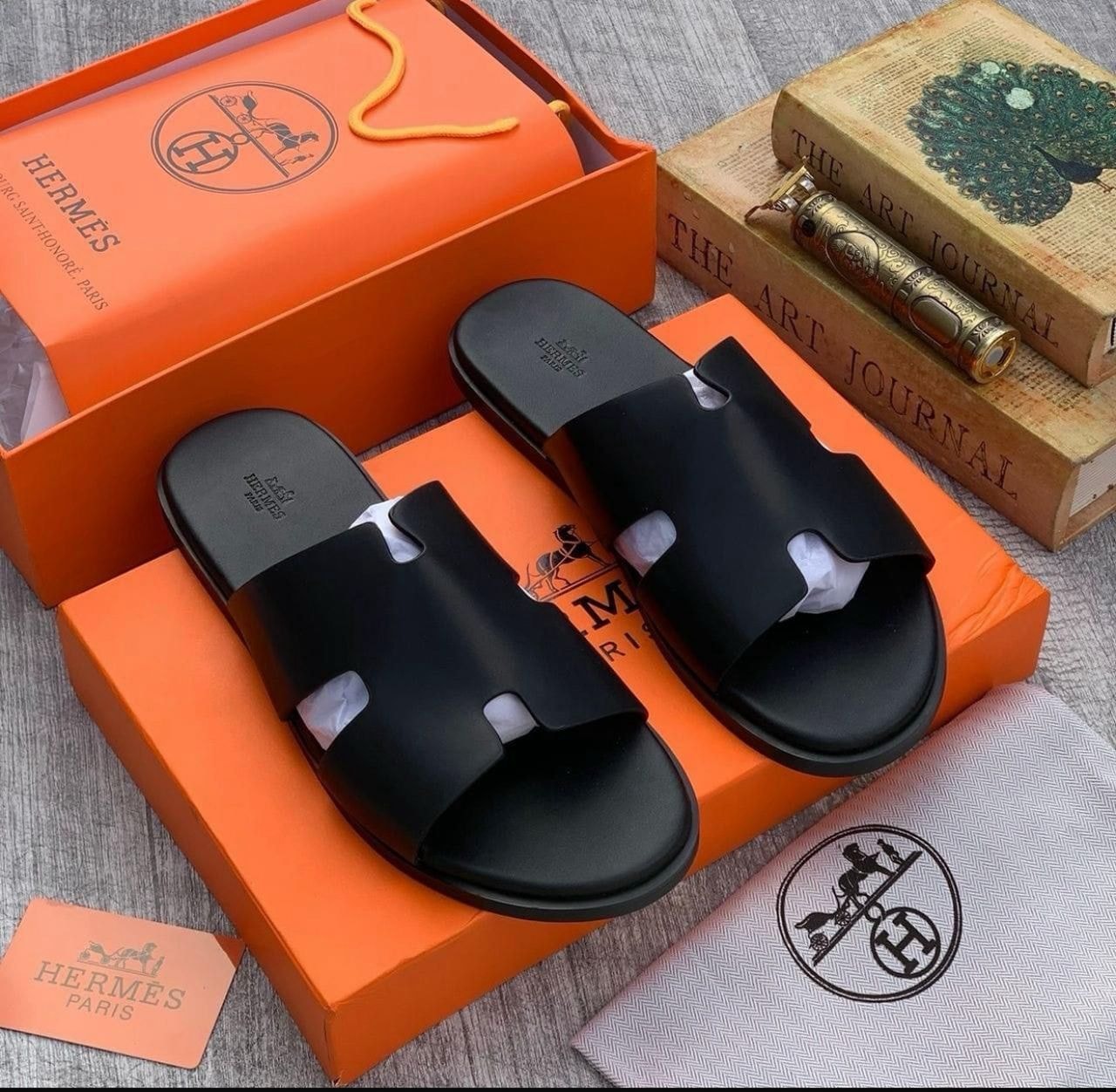 Black Hermes Oran sandals displayed on orange branded boxes with dust bag and accessories.