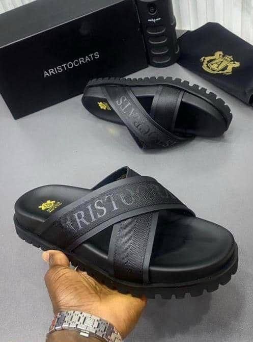 Black Aristocrats slides with cross straps and branding held in hand with matching box and pouch.