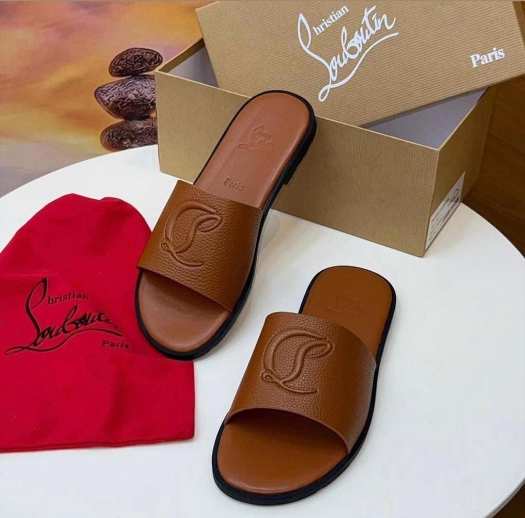 Brown Christian Louboutin slides with logo displayed with branded box, red dust bag and stones on white surface.