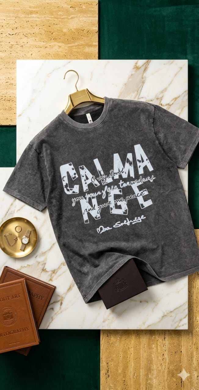 Stonewashed gray t-shirt with CALMA NGE text and cursive quote, displayed on marble with gold tray, watch and books.