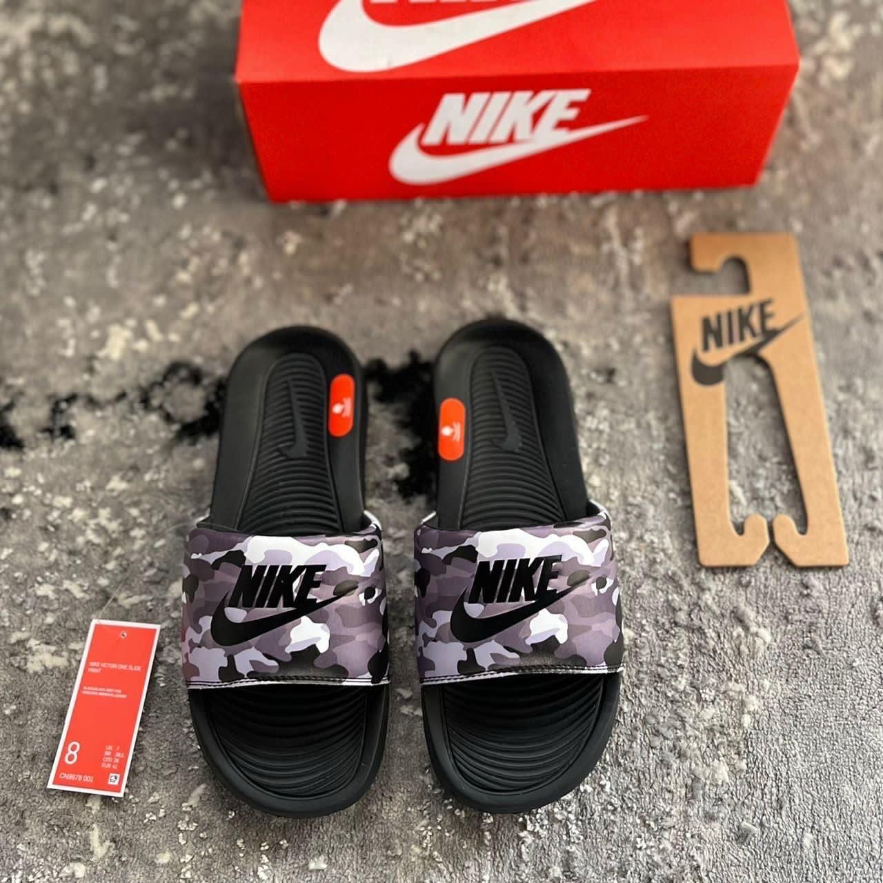 Nike camo slide worn on feet with sole view, red Nike box and decorative items on wooden floor.
