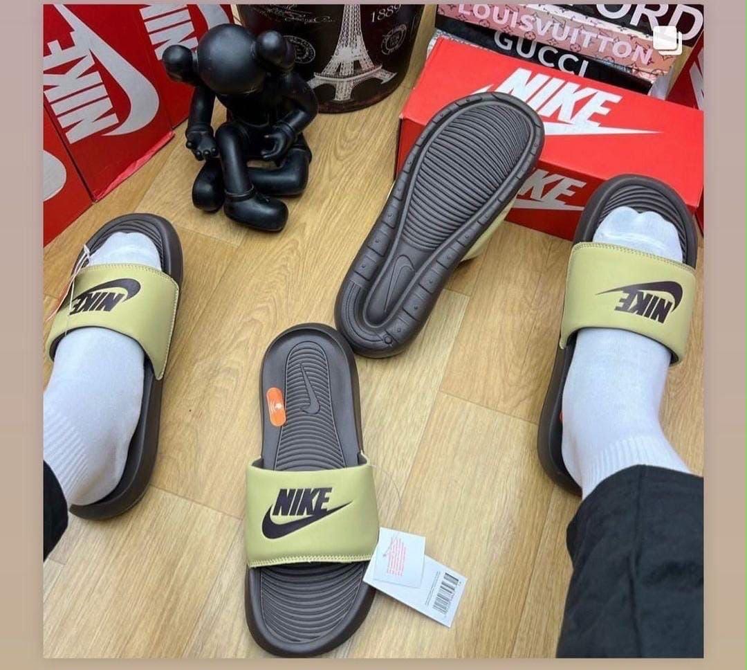 Khaki Nike Victori One Slide worn on feet with sole view on wooden floor.