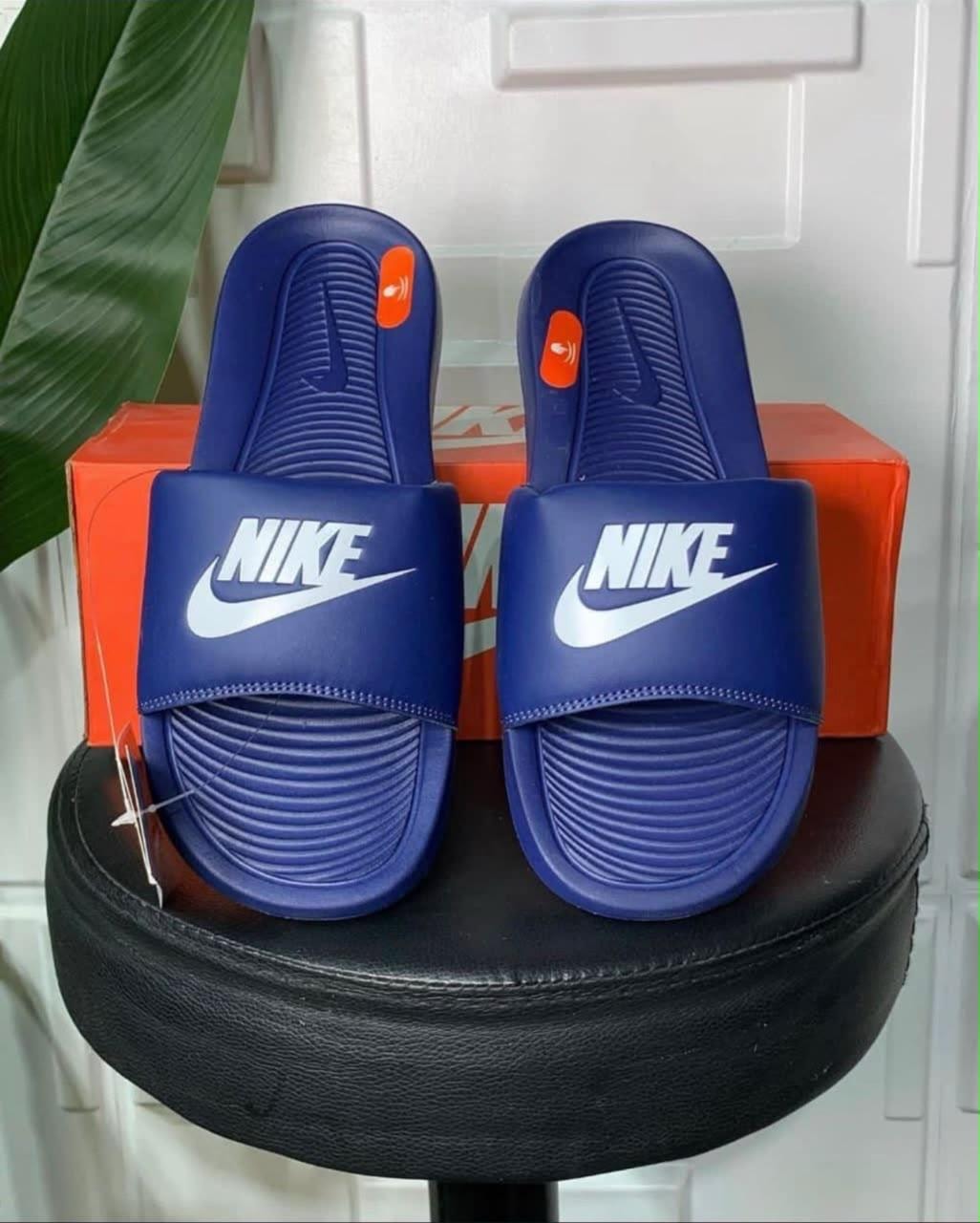 Blue Nike Victori One Slide pair displayed on black stool with green plant.