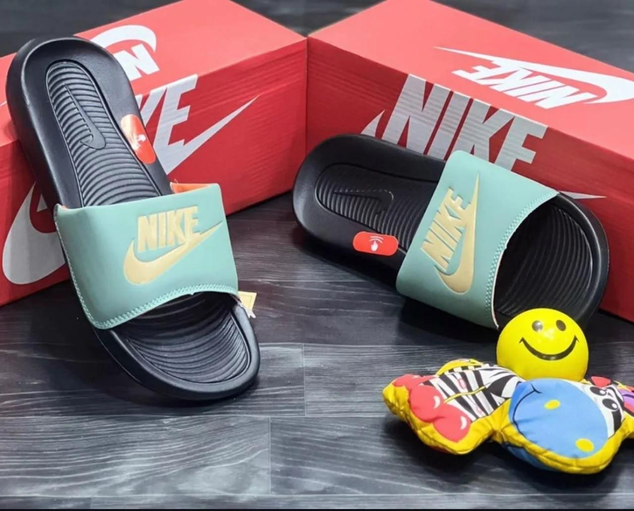 Teal Nike Victori One Slide with gold Swoosh next to red Nike boxes and smiley toy.