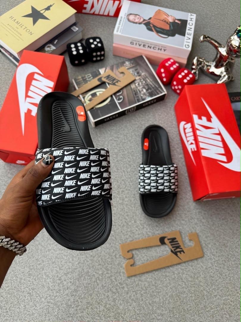 Hand holding black Nike slide with repeating NIKE logo print and second patterned slide on floor with Nike boxes and accessories.