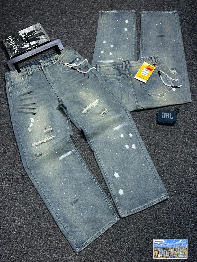 Distressed gray baggy jeans with rips, paint splatters, and beaded chain accessory, flat lay view.