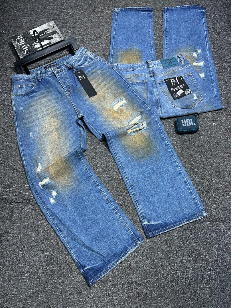 Multiple views of blue baggy distressed jeans with rips, yellow fading, tags and lifestyle items on gray surface.