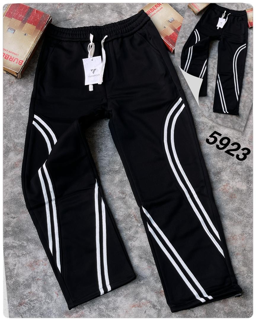 Black joggers with white curved side stripes and TXFAMOUS tag on gray surface with boxes.