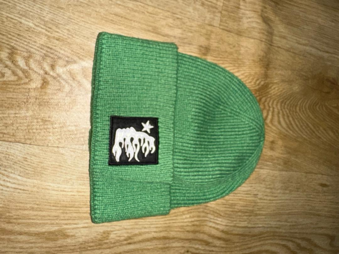 Green knit beanie with black patch logo on wooden surface.
