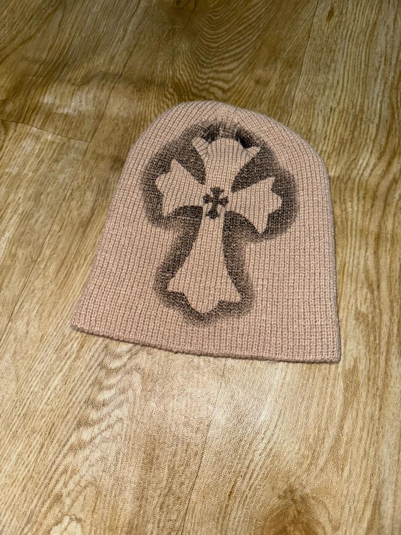 Beige knit beanie with large dark cross design on wooden floor.
