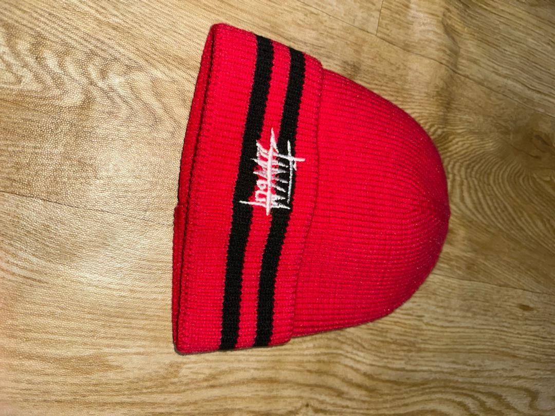 Red knit beanie with black stripes and white Stussy logo on wooden surface.