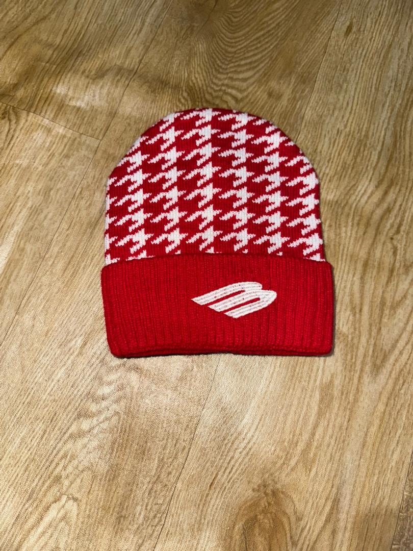 Red and white houndstooth knit beanie with logo on cuff laid on wooden surface.