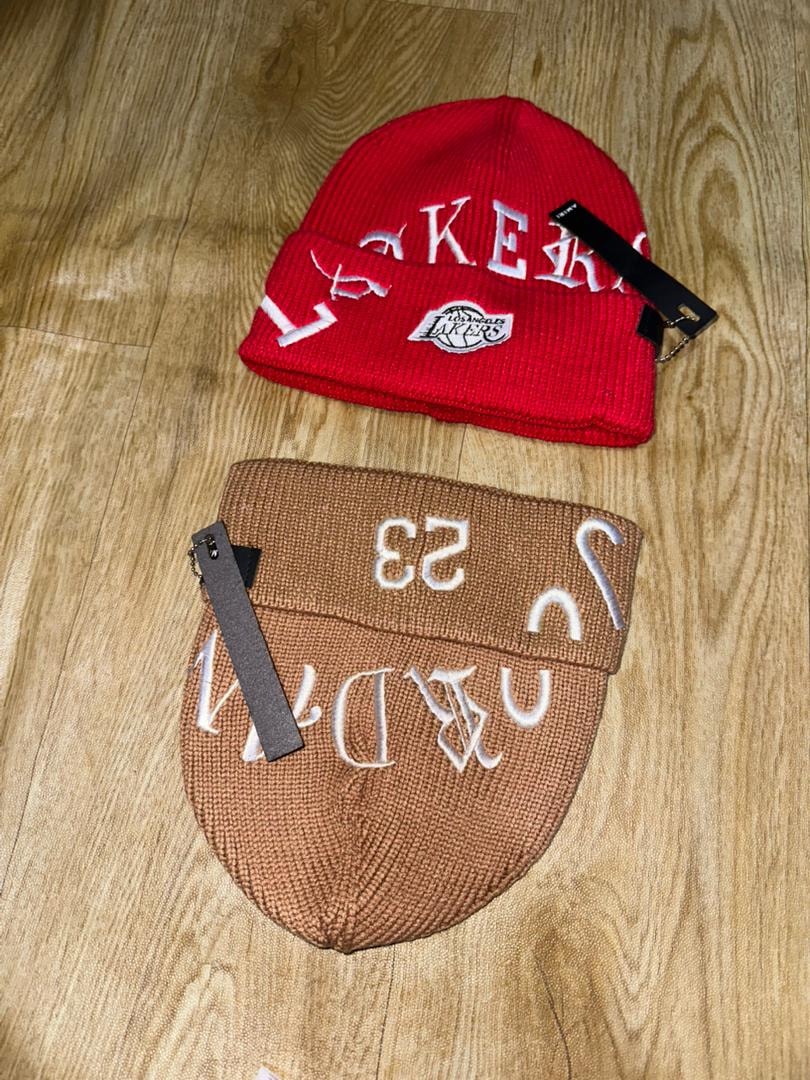 Brown embroidered beanie with white 23 and designs next to red Lakers beanie on wooden floor.
