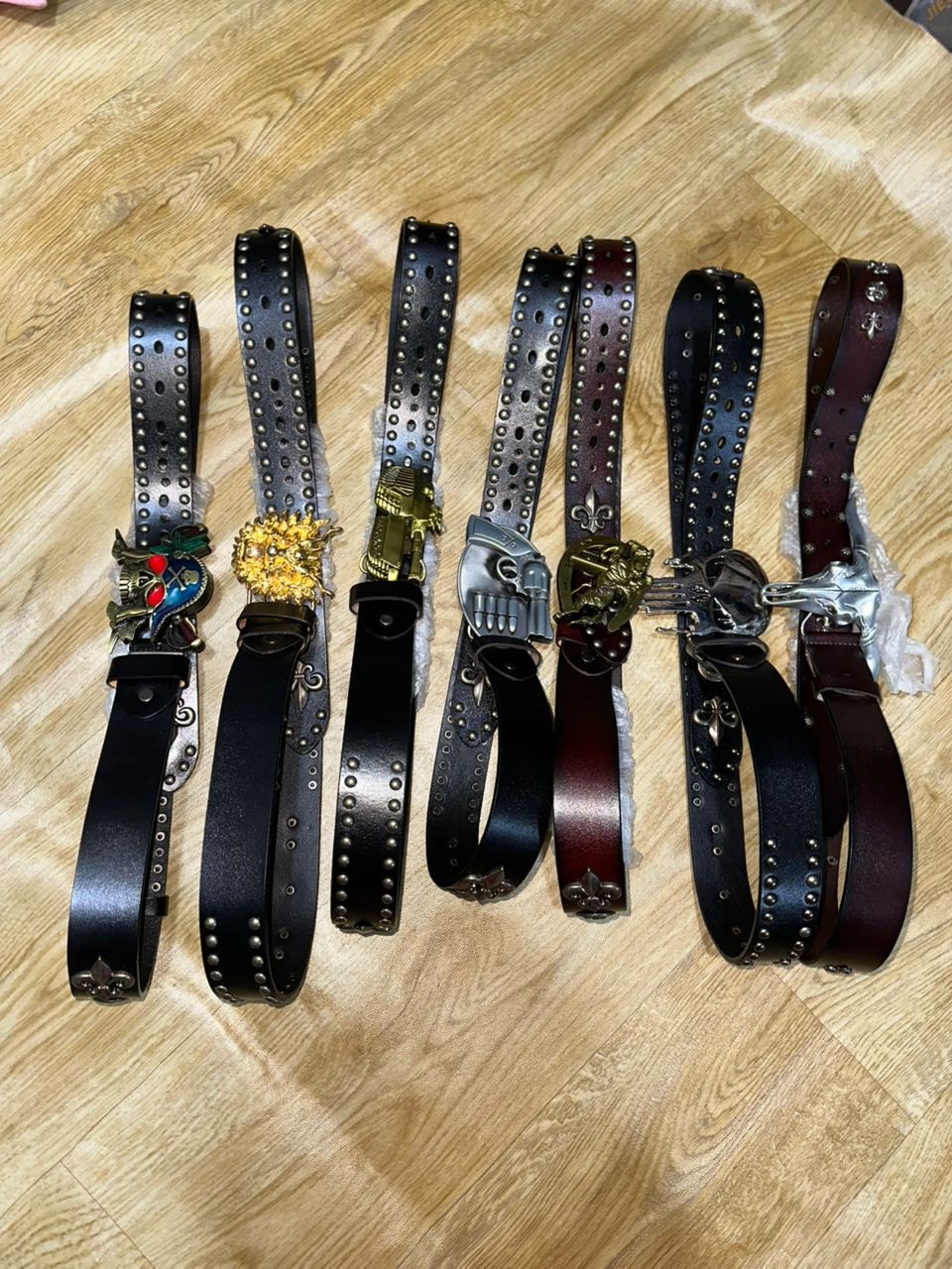 Six studded leather belts with decorative metal buckles in black and burgundy on wooden floor.