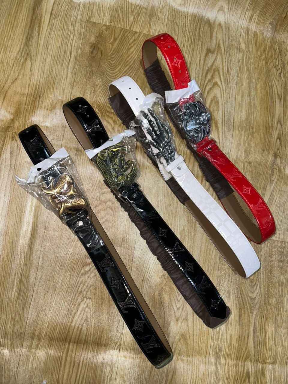 Four monogram embossed belts in black, white and red with ornate buckles on wooden surface.