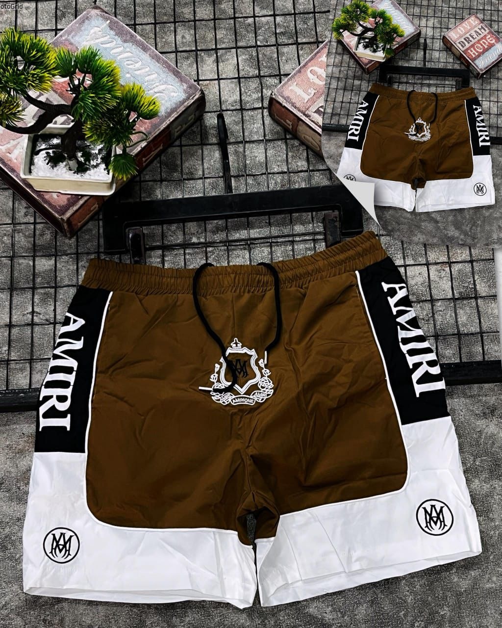 Brown Amiri logo shorts with crest emblem and side text styled on grid with props.