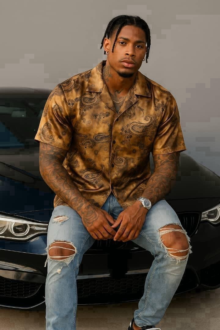 Man seated on car wearing brown paisley pattern short sleeve shirt with jeans.