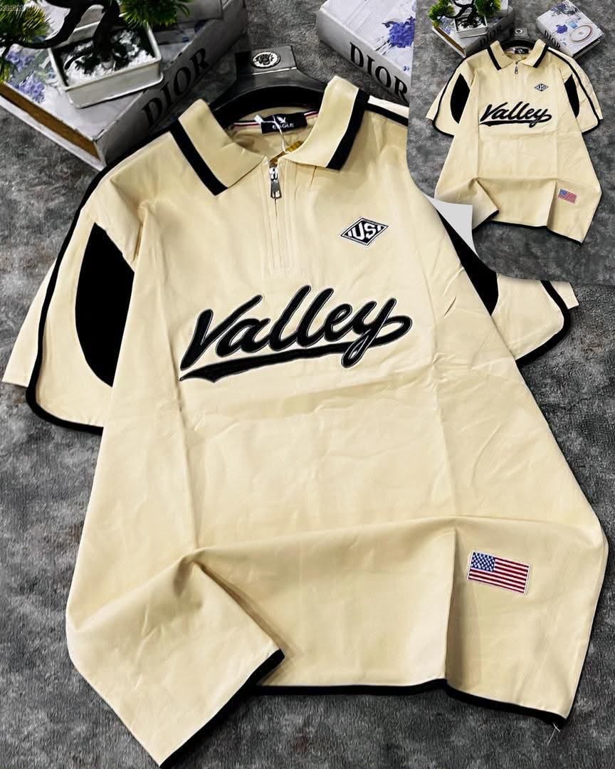 Cream Valley script polo shirt laid flat on gray surface showing front embroidery and black contrast panels.