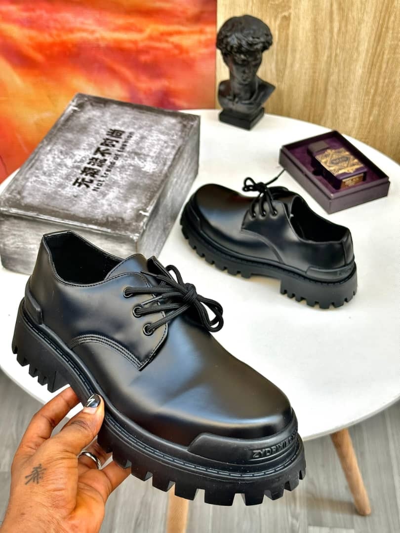Black chunky lace-up derby shoes held by hand with grey box and accessories.