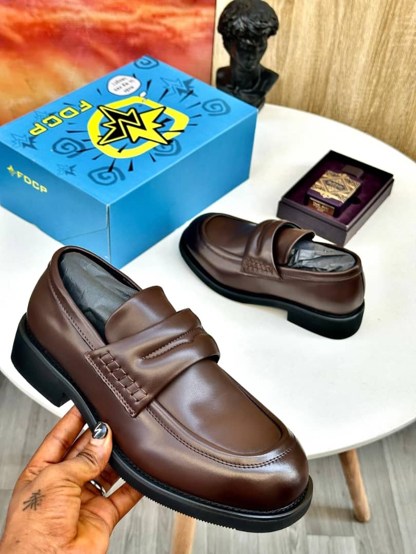 Brown FDCP penny loafers held by hand with blue box and bust in background.