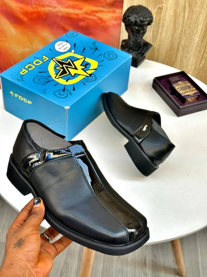 Black FDCP buckle loafers held by hand with blue box and accessories in background.