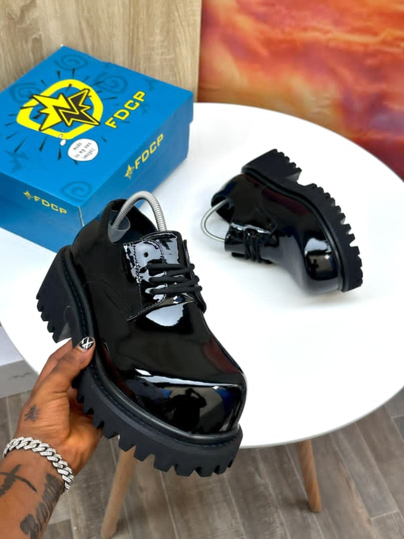 Black patent platform derby shoes held in hand with blue FDCP box in background.