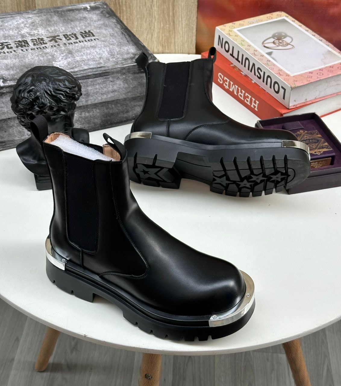 Black Chelsea boots with metal details displayed on white table with decorative boxes and bust.