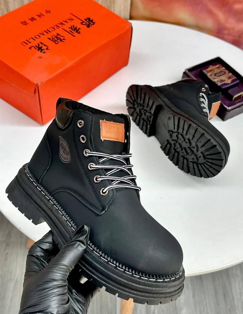 Black chunky ankle boots held in gloved hand showing side profile and sole with orange product box.