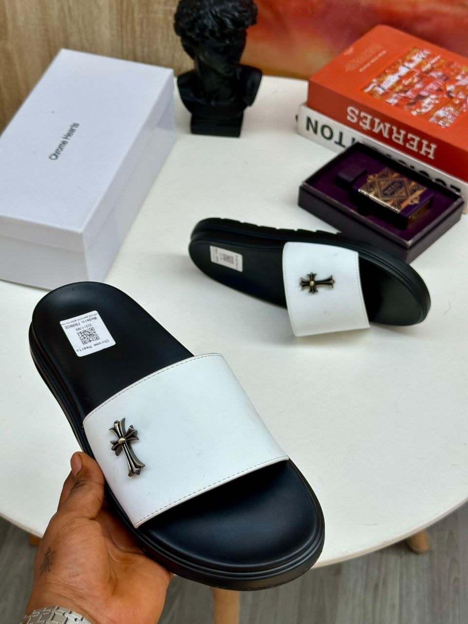 White and black Chrome Hearts slides with cross emblem held in hand.