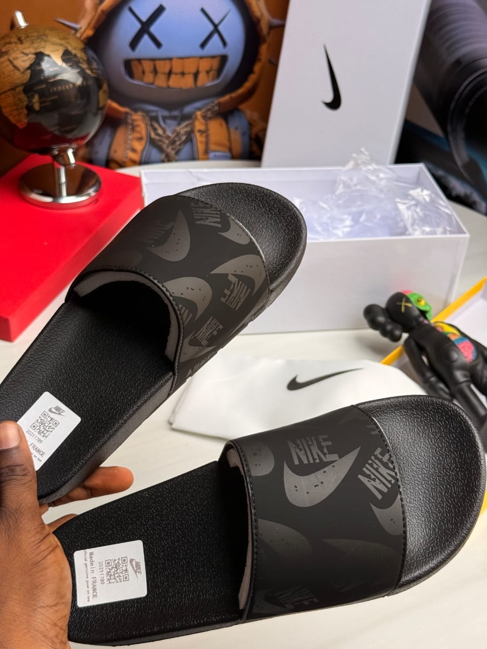 Black Nike slides with repeating swoosh pattern held in hand with box.