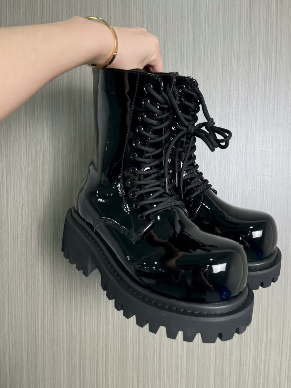 Black patent leather combat boots held by hand against gray wall.