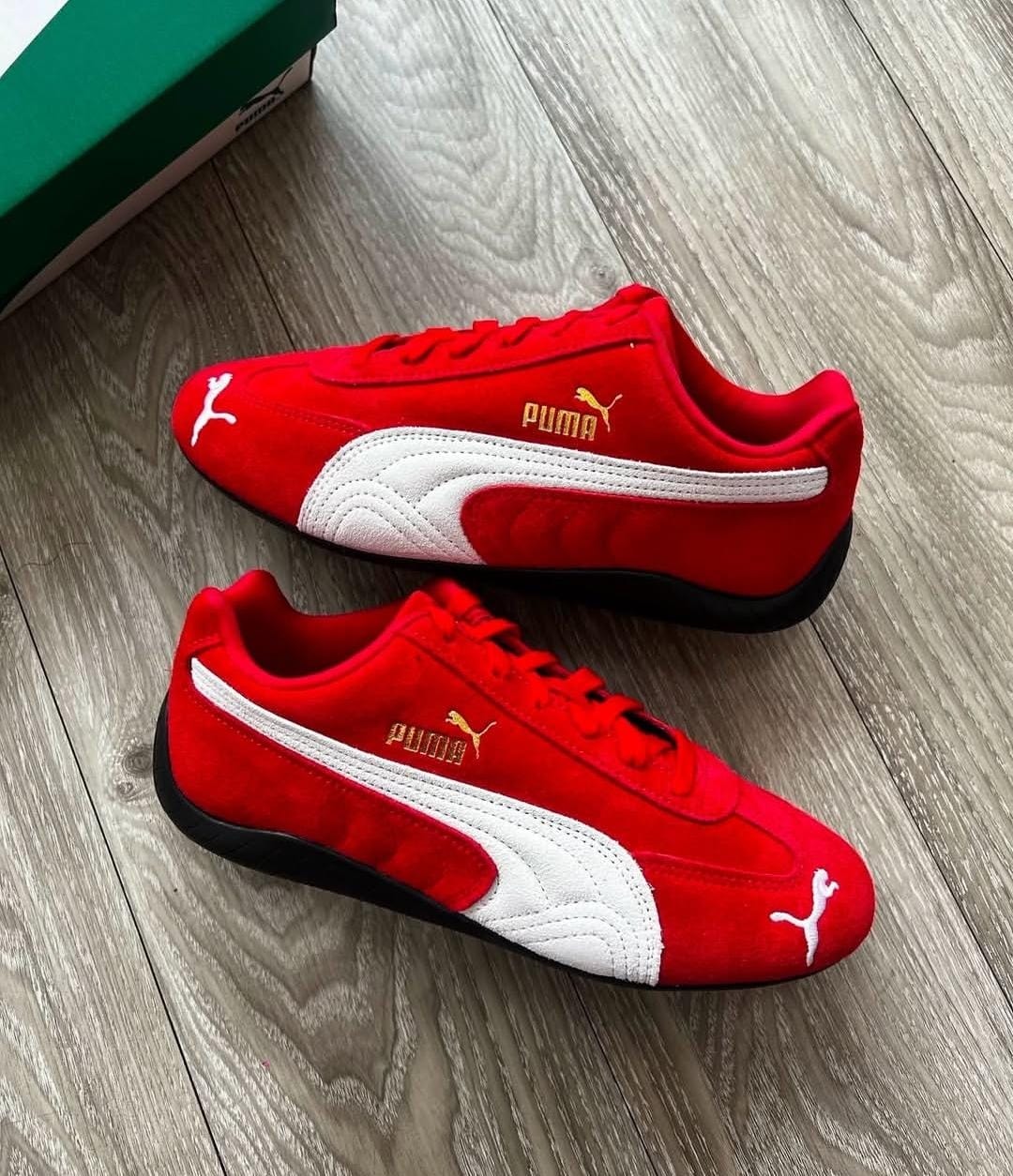 Red Puma suede sneakers with white stripe and gold branding on wooden floor.