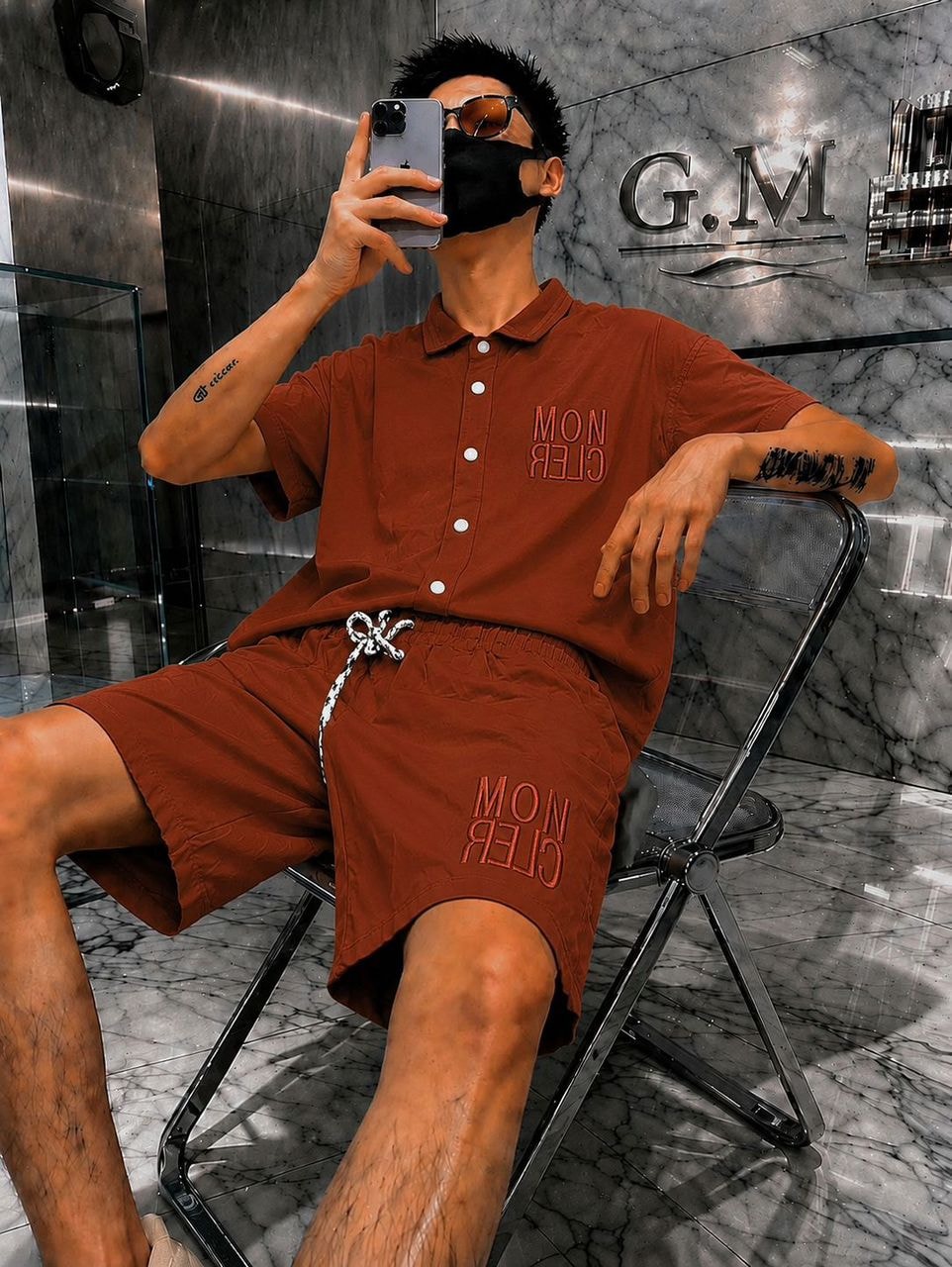 Man in brown polo shirt and embroidered shorts with Moncler logo, seated and taking a mirror selfie.