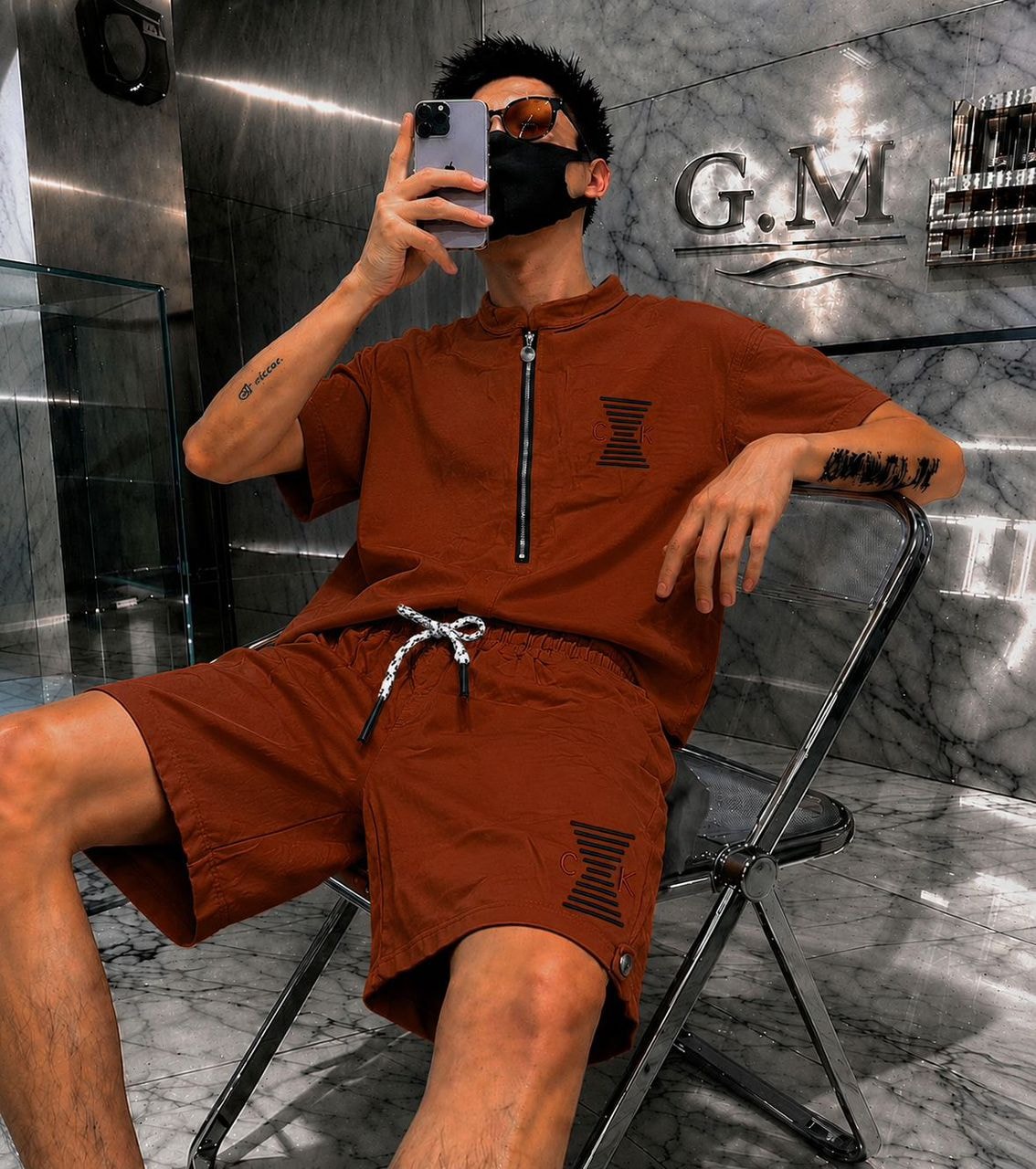 Man wearing brown zip-neck top and shorts with CK logos, seated in luxury marble setting.