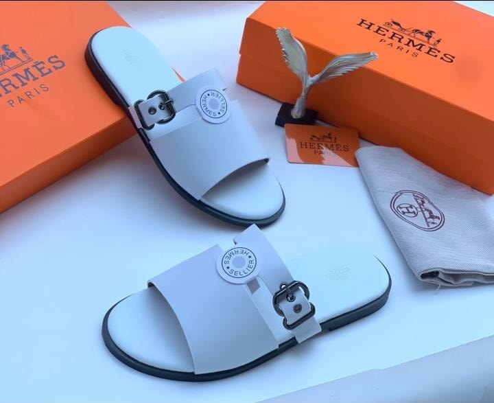 Hermes white slide sandals with buckle and circular tag alongside orange boxes and accessories.