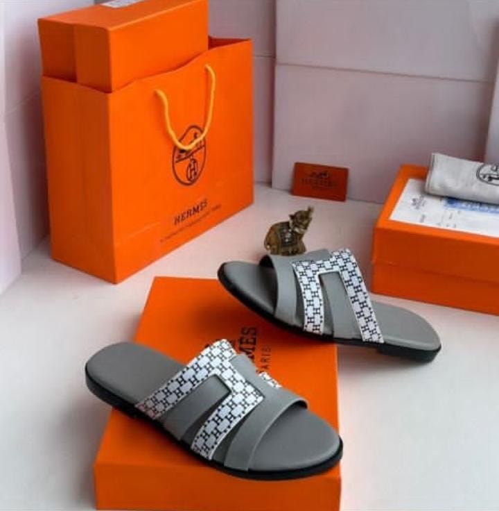 Hermes gray slide sandals with patterned H straps on orange luxury boxes.