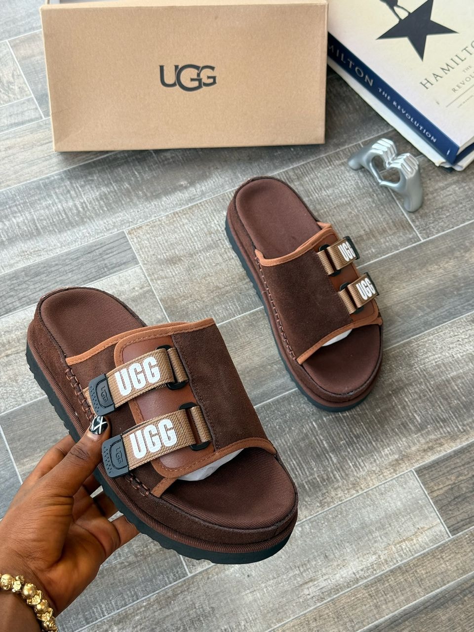 Brown UGG slides with logo straps held by hand next to box on tiled floor.