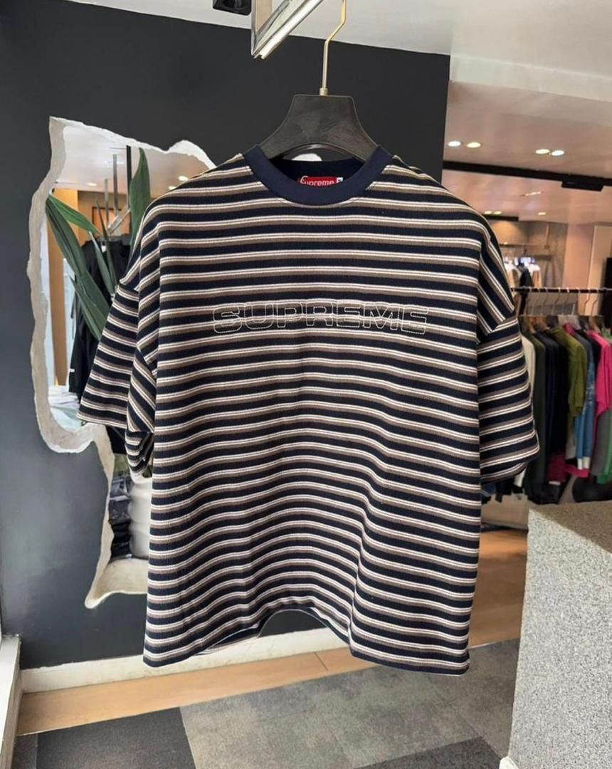 Brown and white striped Supreme t-shirt hanging in store setting with SUPREME logo visible.