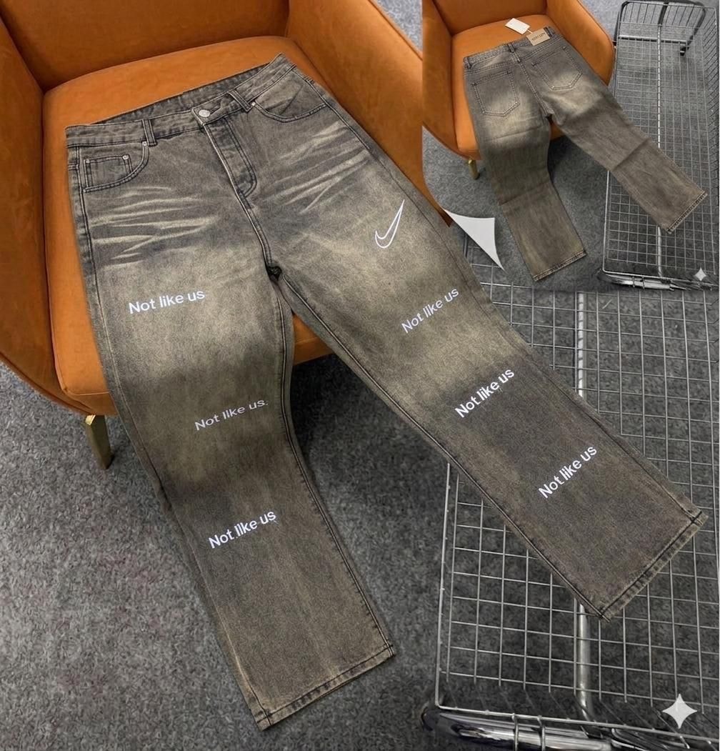 Gray jeans with repeated white "Not like us" text prints and Nike swoosh logo on the leg.